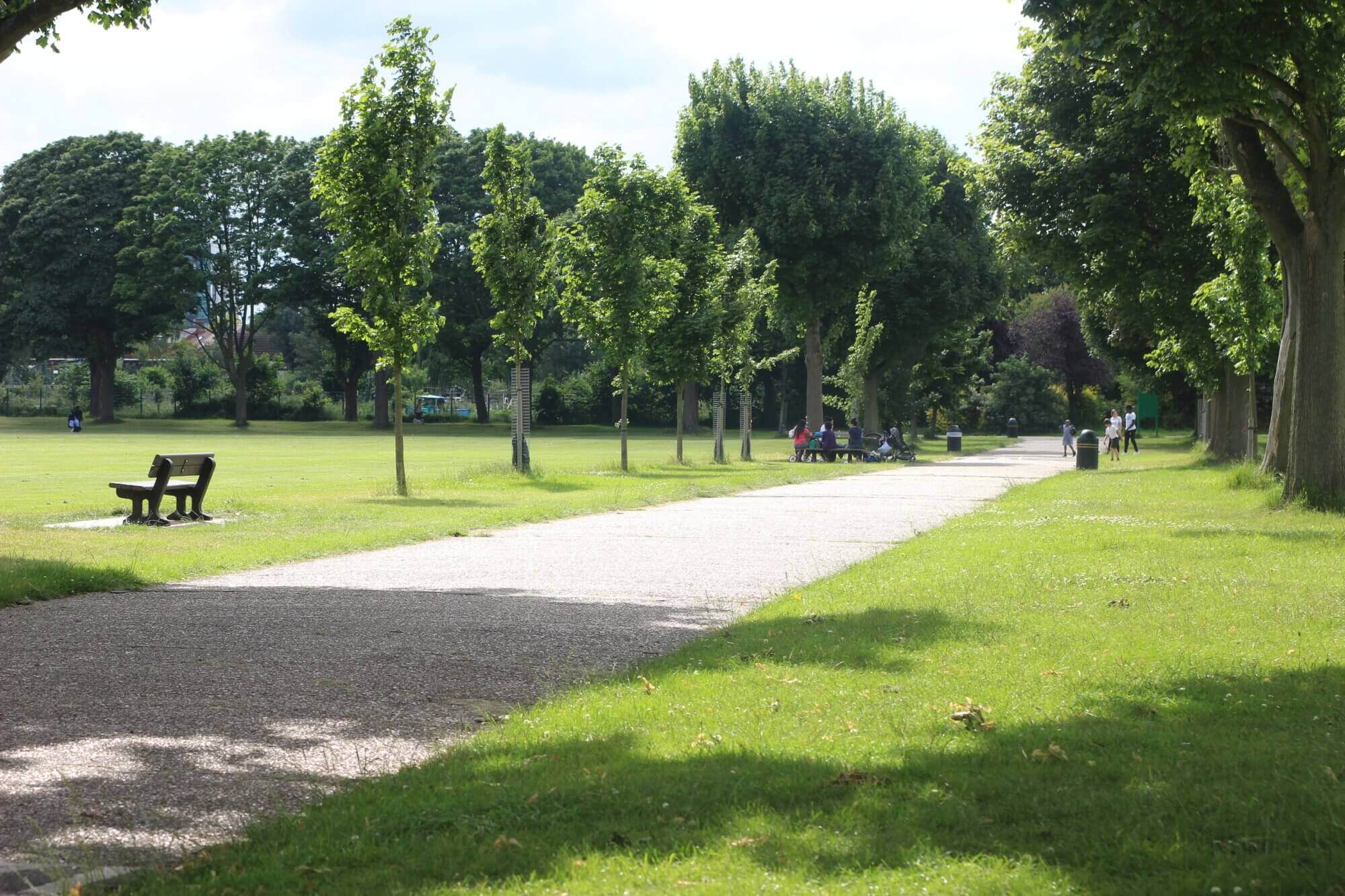 A paved path lined with trees in a park, with people sitting on benches and walking in the distance. - Home Instead