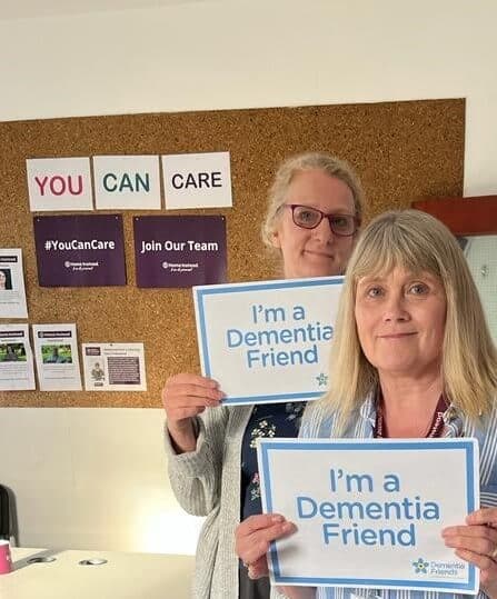 Two people holding signs that say "I'm a Dementia Friend" in an office with supportive posters on the wall. - Home Instead