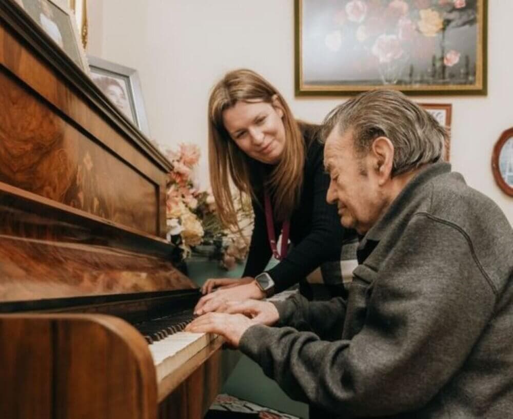 Elderly man playing piano with assistance from a woman in a cozy room with floral paintings on the wall. - Home Instead