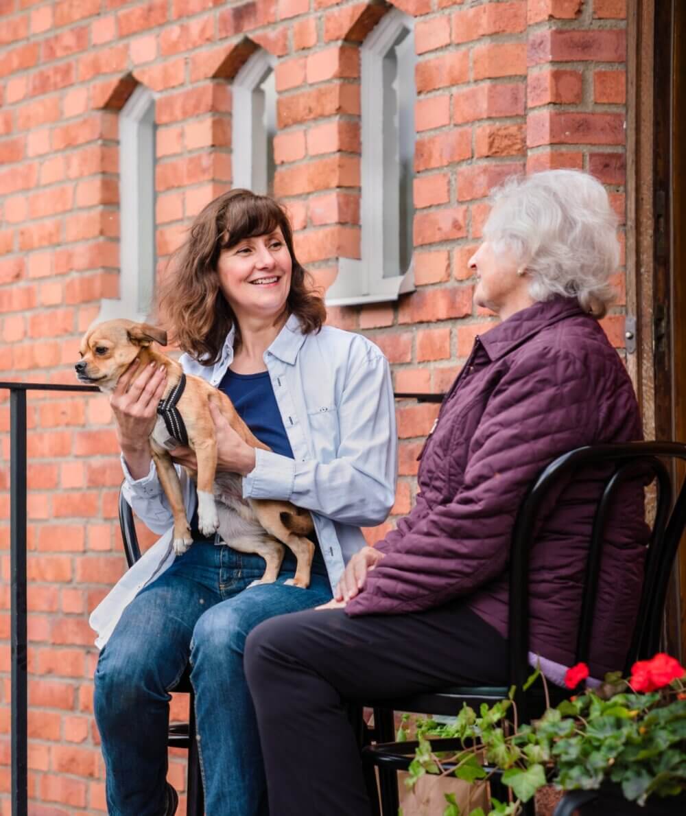 Two women sitting on chairs outside a brick building, one holding a small dog and both smiling at each other. - Home Instead