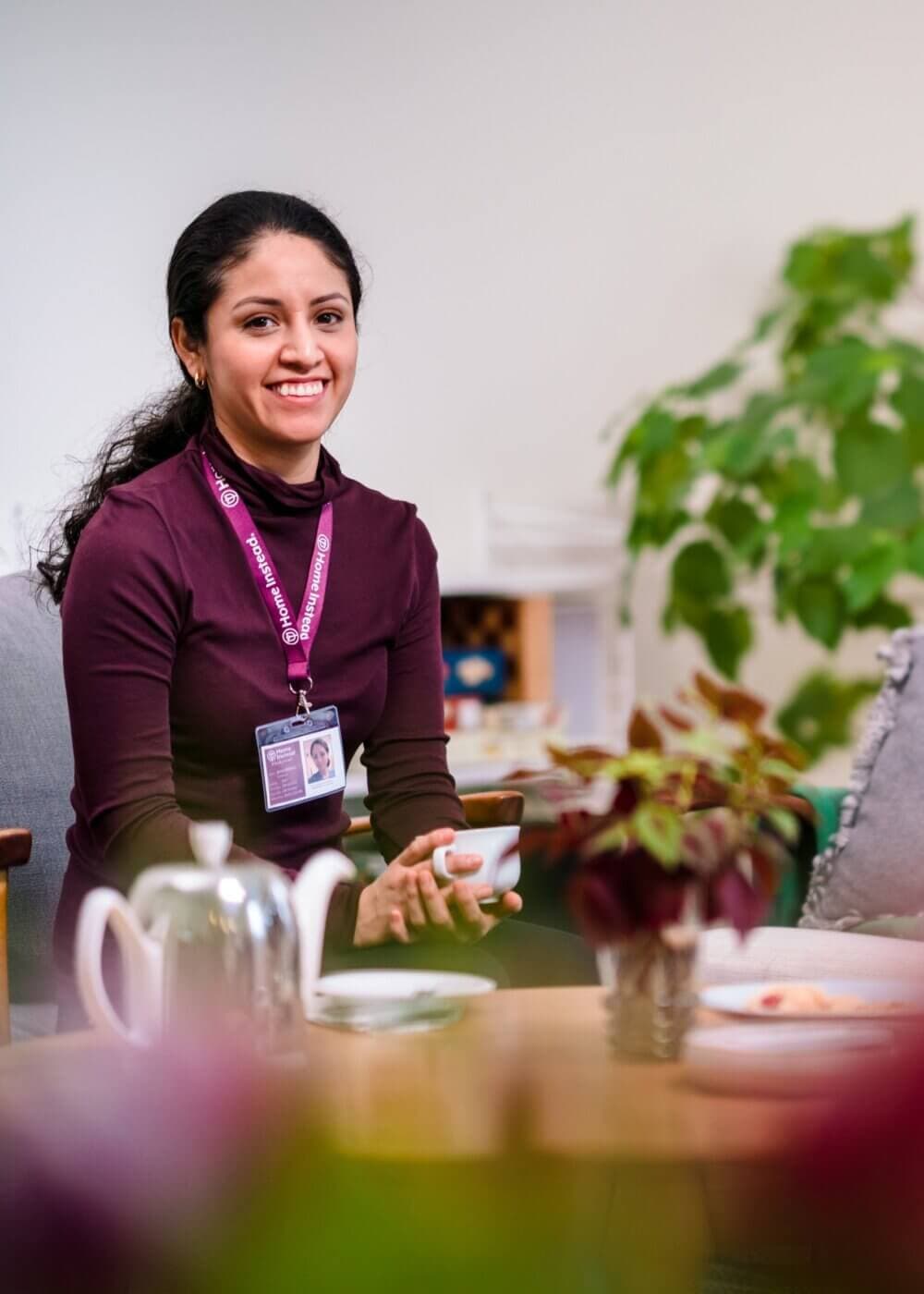 A woman in a maroon top, wearing a badge, sits at a table with a teapot, smiling. - Home Instead