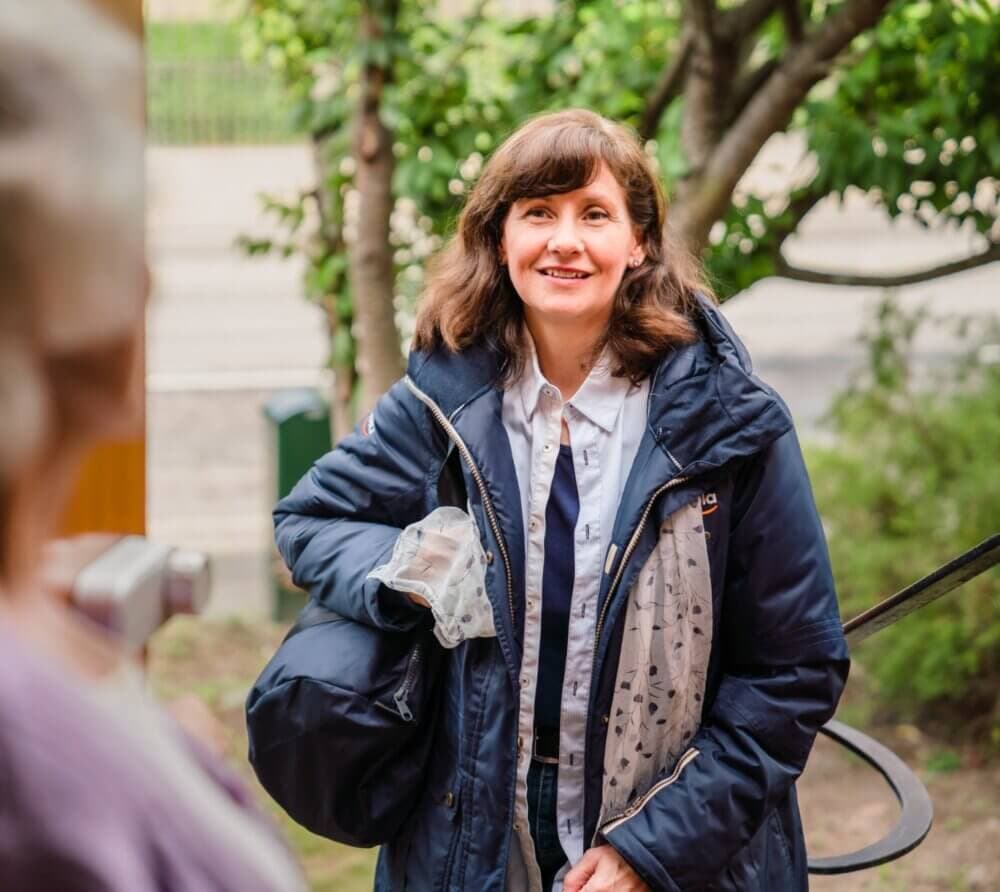 A woman in a jacket smiles while standing outside near trees, holding bags and looking at another person. - Home Instead