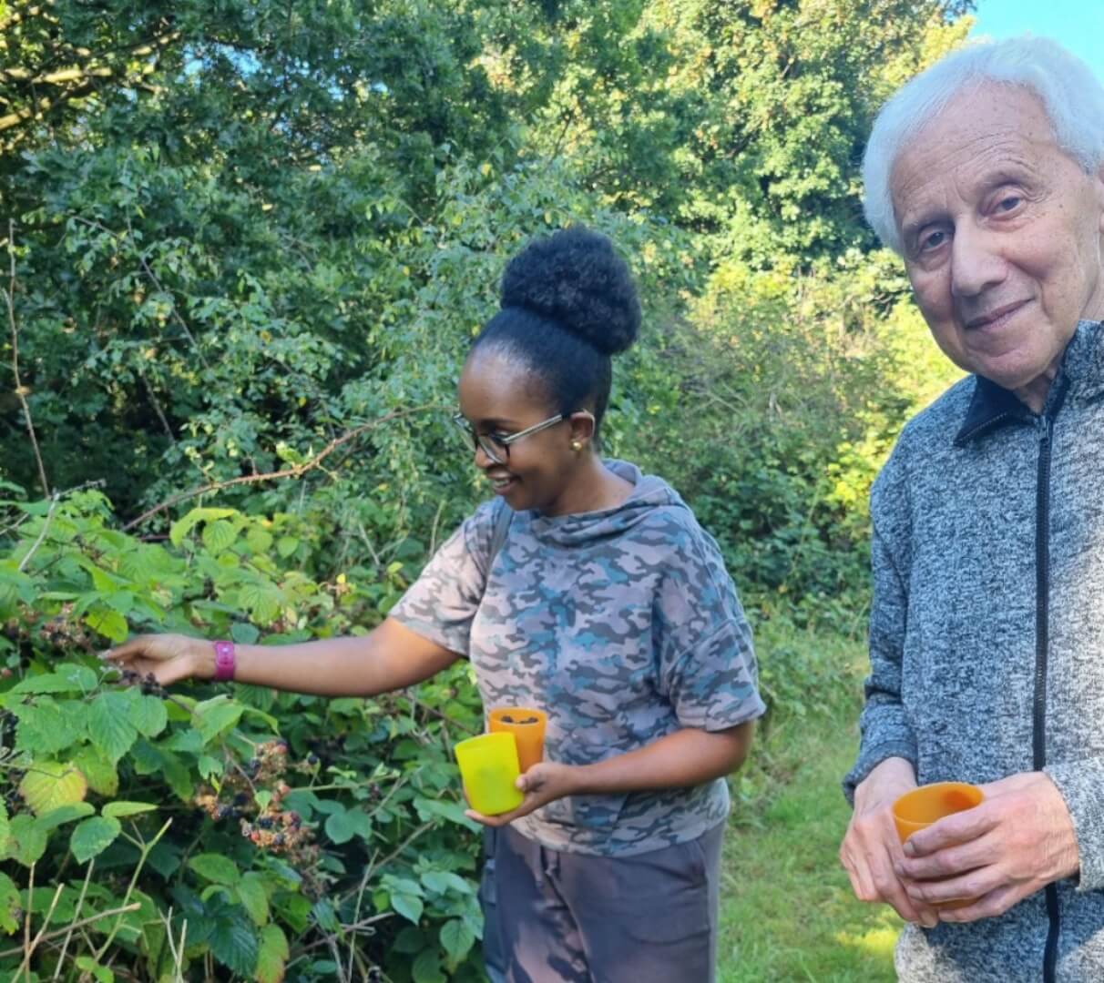 Two people standing outdoors picking berries, both holding yellow containers, surrounded by green foliage. - Home Instead