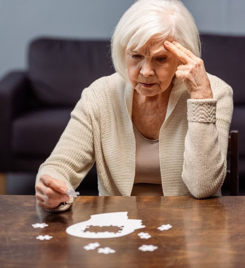 Elderly woman concentrating on assembling a jigsaw puzzle at a wooden table in a cozy, dimly lit room. - Home Instead