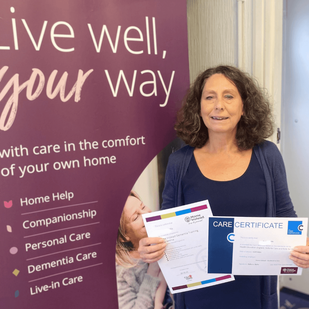 Woman standing with two certificates in hand, smiling beside a banner that says "Live well, your way" and offers various care services. - Home Instead