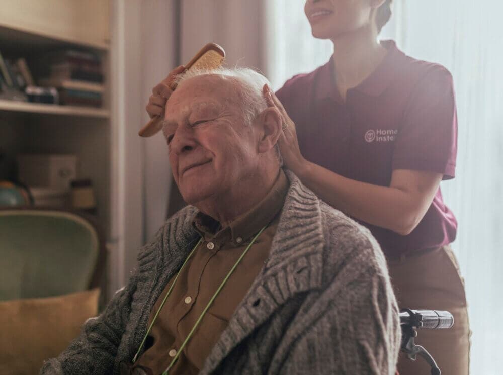 A caregiver brushes the hair of a smiling elderly man in a wheelchair inside a cozy room. - Home Instead