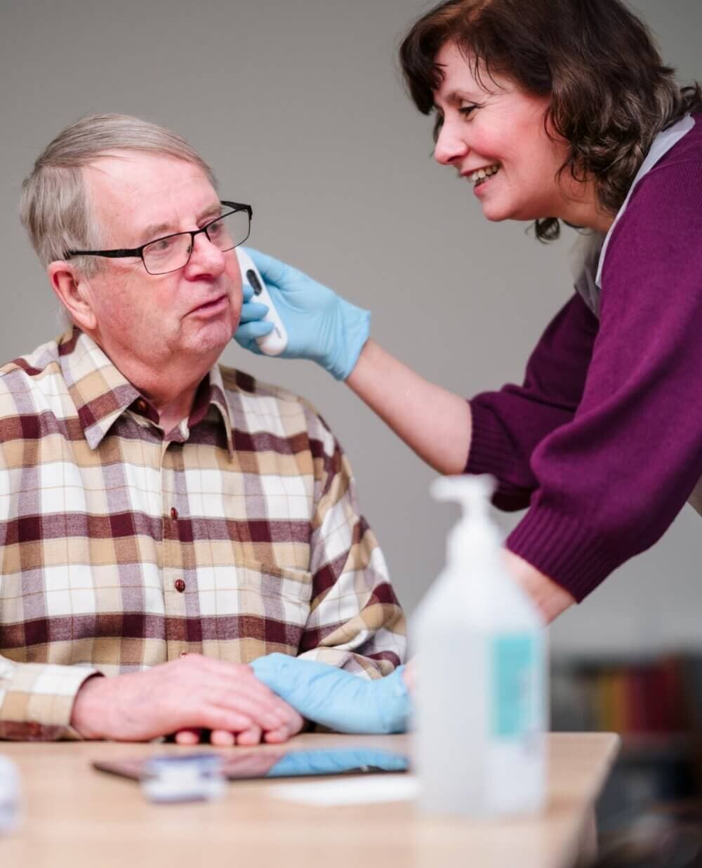 A caregiver wearing gloves helps an elderly man in a plaid shirt with a hygiene task, both smiling warmly. - Home Instead