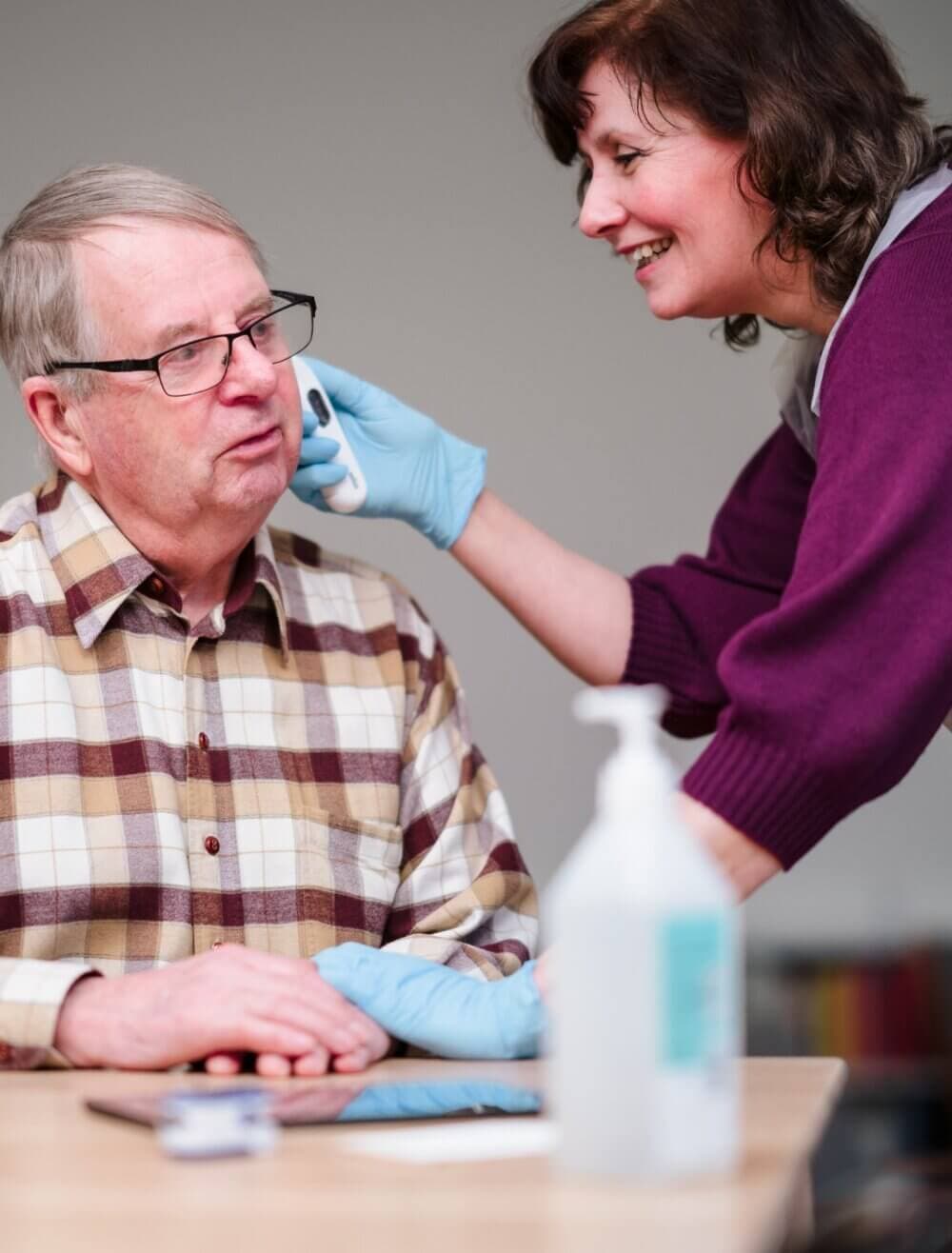 A caregiver in gloves helps an elderly man with a medical task at a table with hand sanitizer. - Home Instead