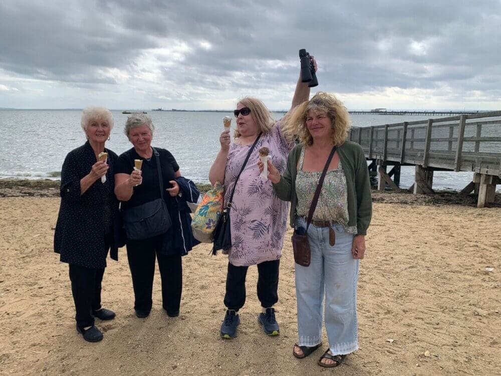 Four women enjoying ice cream while standing on a beach near a wooden pier on a cloudy day. - Home Instead