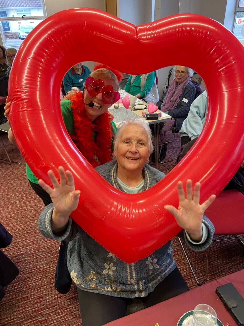 Two women smiling through a large red heart-shaped balloon frame, with a group of people seated in the background. - Home Instead