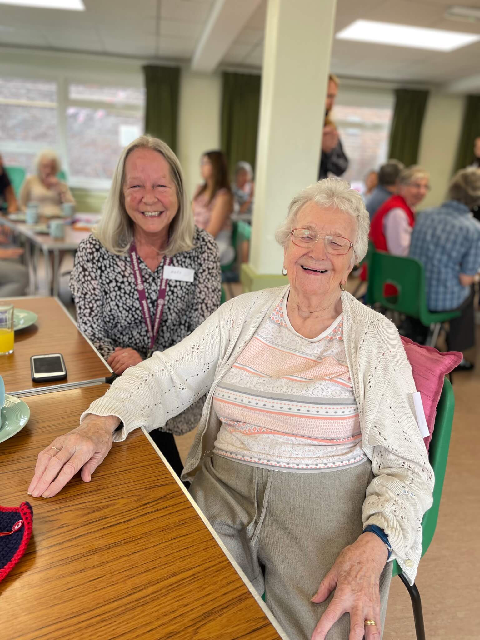 Two elderly women sitting together and smiling at a social gathering, with others seated in the background. - Home Instead