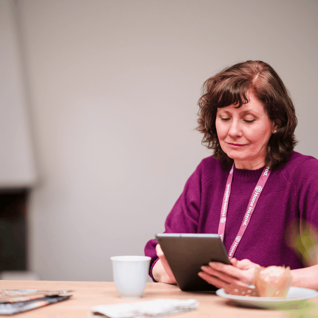 Woman with lanyard sitting at a table, looking at a tablet, with a coffee cup and croissant in the foreground. - Home Instead