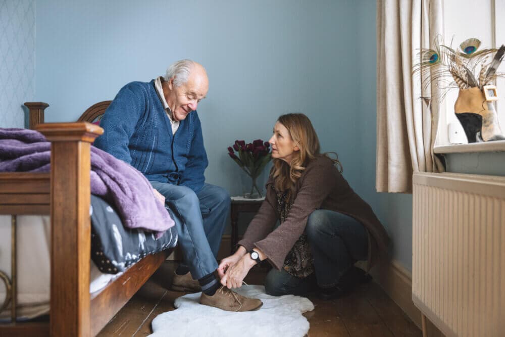 An elderly man sits on a bed while a woman kneels down and helps him put on a shoe in a cozy room. - Home Instead