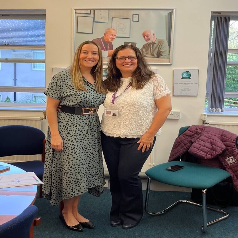 Two women stand smiling together in an office in front of a portrait of two men, with chairs and coats visible around them. - Home Instead