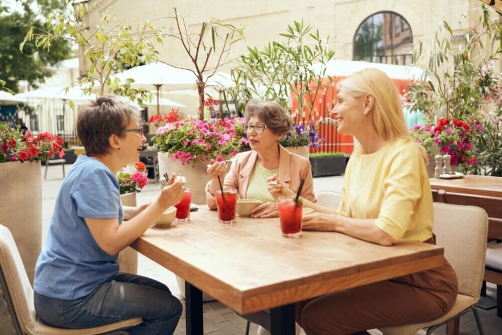 Three women enjoy drinks and conversation at an outdoor cafe on a sunny day, surrounded by colorful flowers. - Home Instead