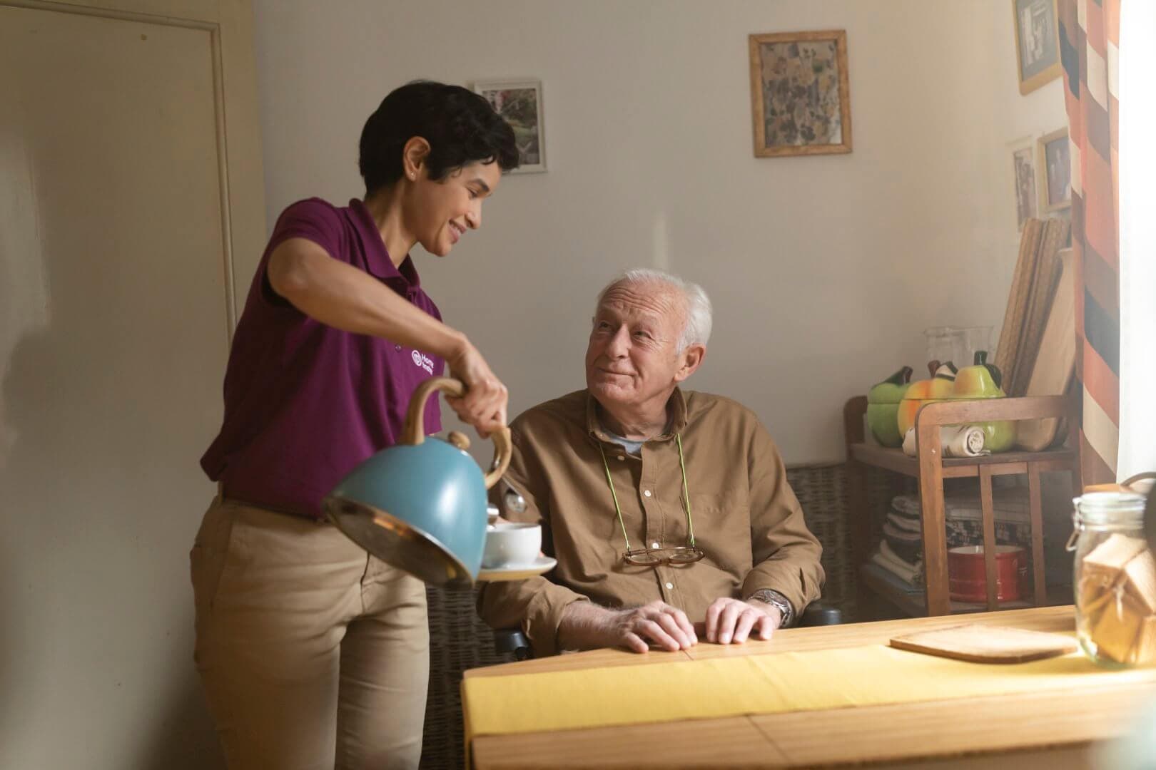 A caregiver pours tea for an elderly man sitting at a table in a cozy room with framed photos on the wall. - Home Instead