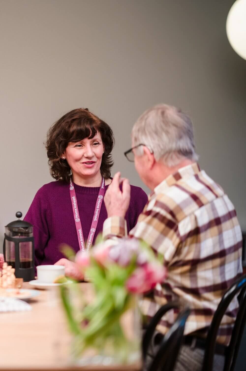 Two people sitting at a table, engaged in conversation. A French press and a vase of flowers are in the foreground. - Home Instead