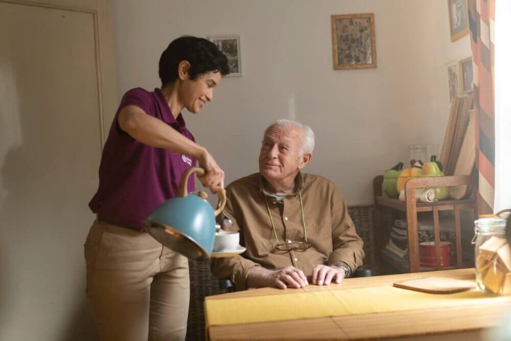 A woman in a purple sweater pours coffee for an elderly man sitting at a table with a flower vase. - Home Instead