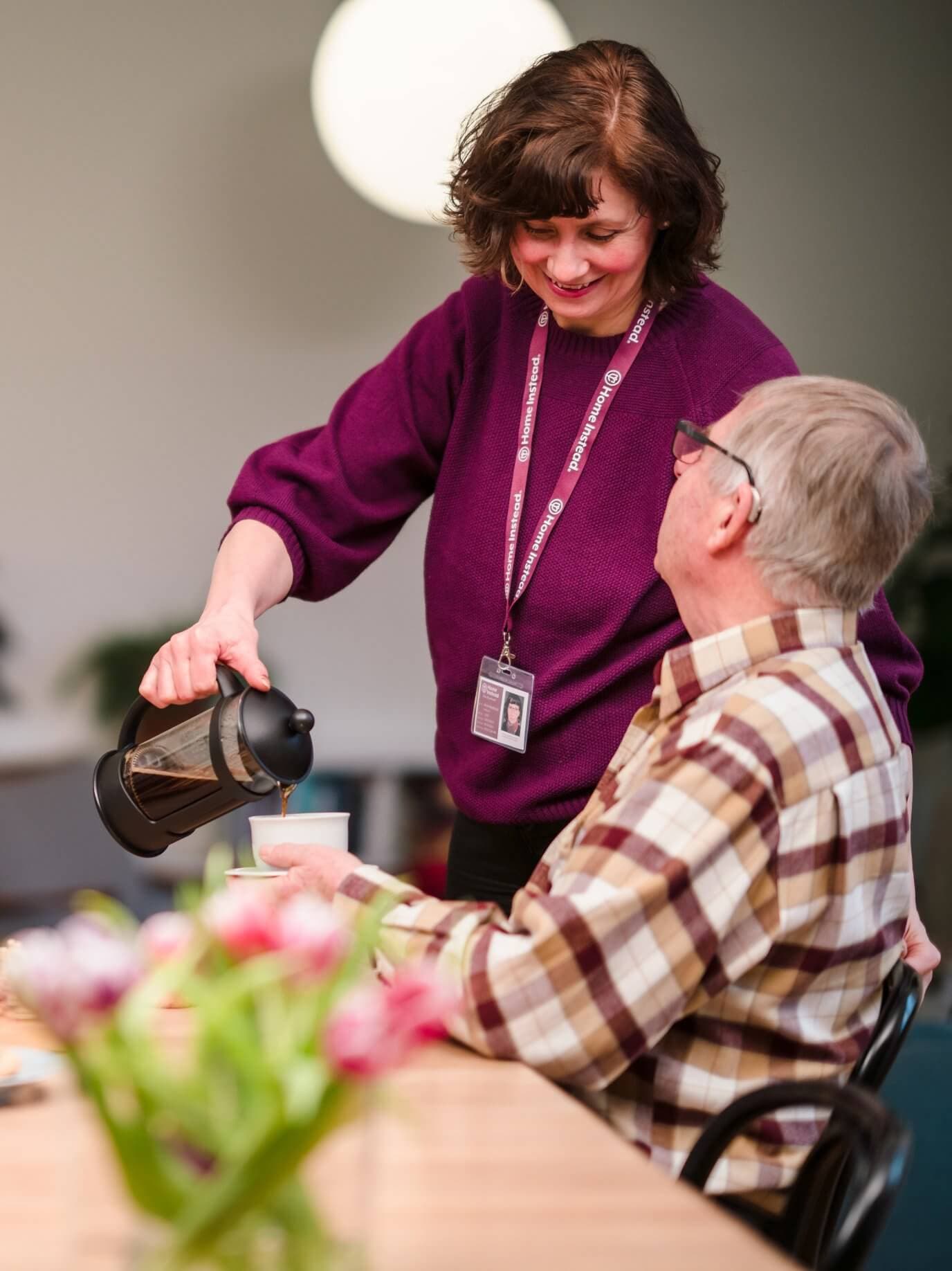 A woman in a purple sweater pours coffee for an elderly man sitting at a table with a flower vase. - Home Instead