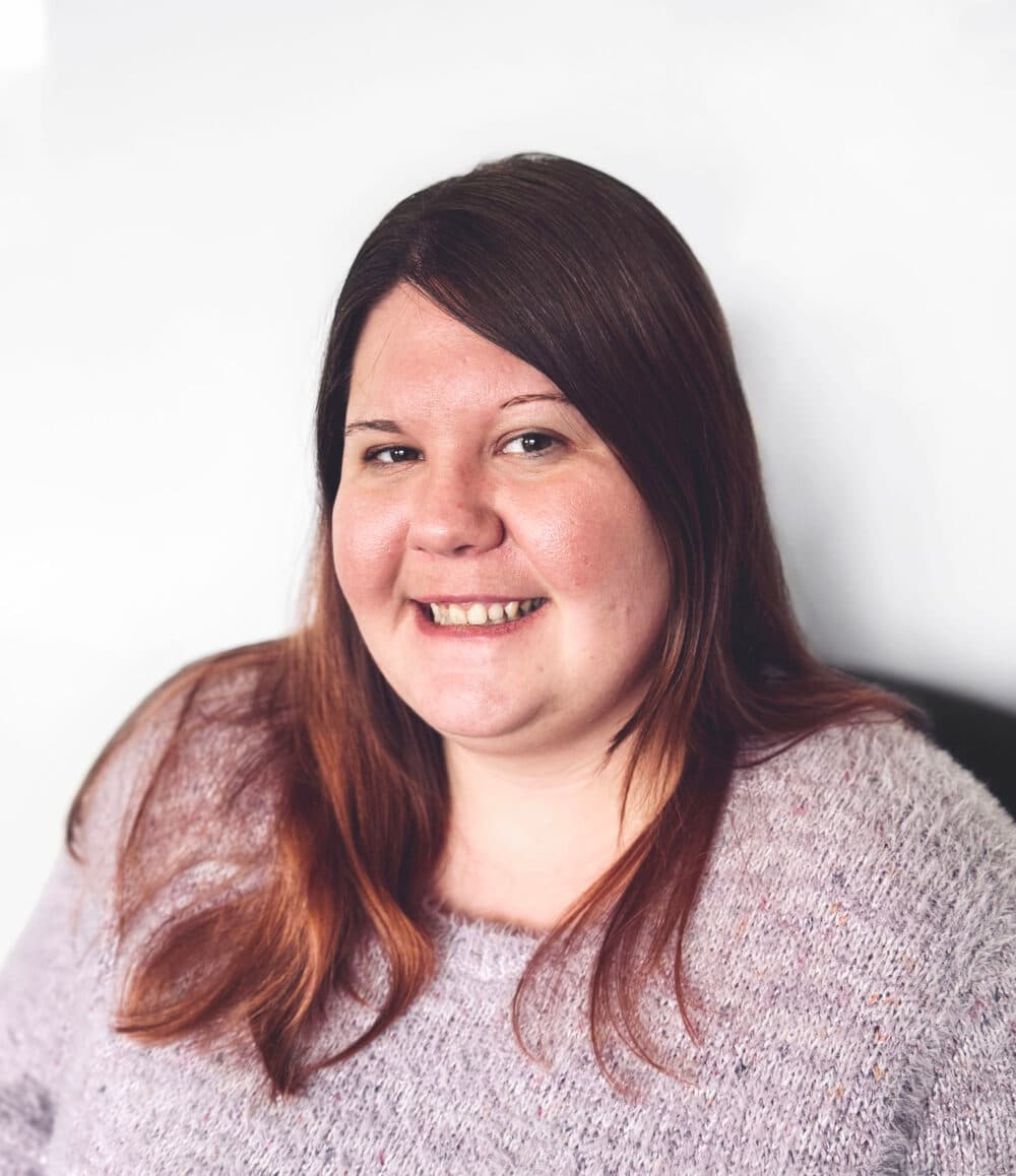 A woman with long brown hair, wearing a light purple sweater, smiles at the camera against a plain background. - Home Instead