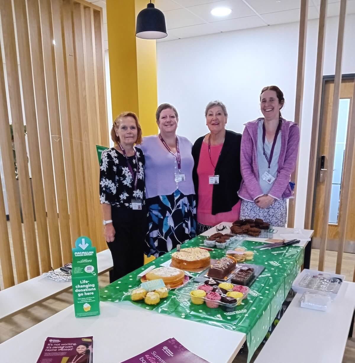 Four women standing behind a table with a variety of baked goods in a brightly lit room, smiling at the camera. - Home Instead Poole
