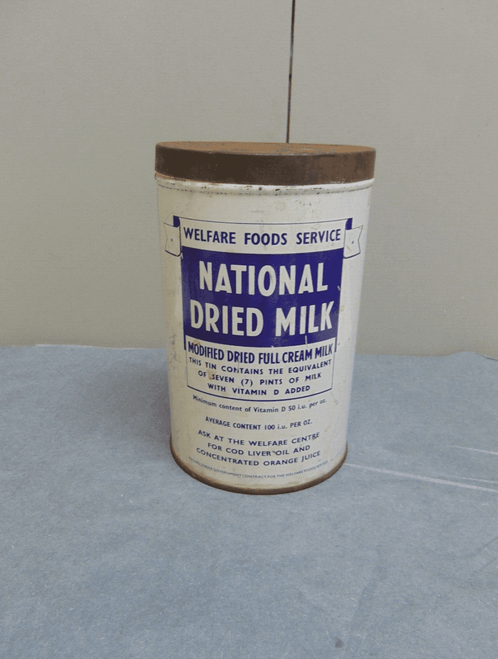 A vintage tin labeled "National Dried Milk" from the Welfare Foods Service, placed on a blue cloth background. - Home Instead