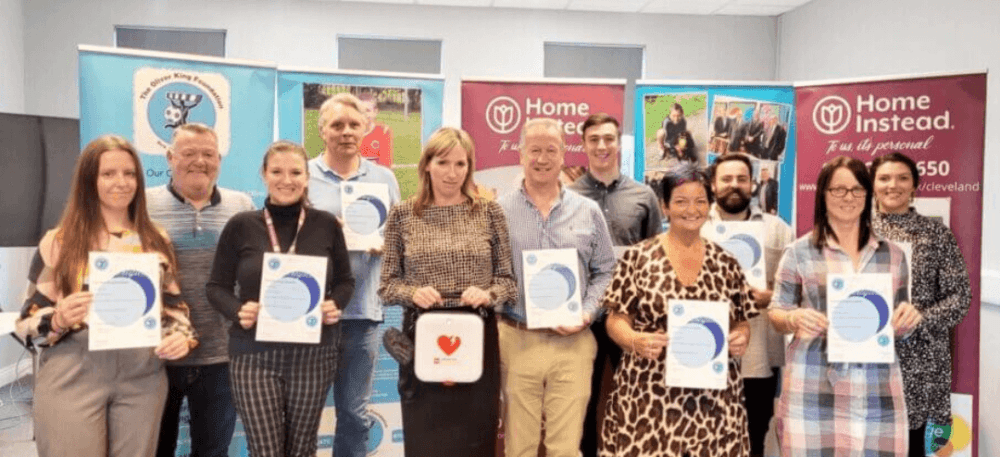 A group of people proudly holding certificates standing in front of colorful banners in a room. - Home Instead