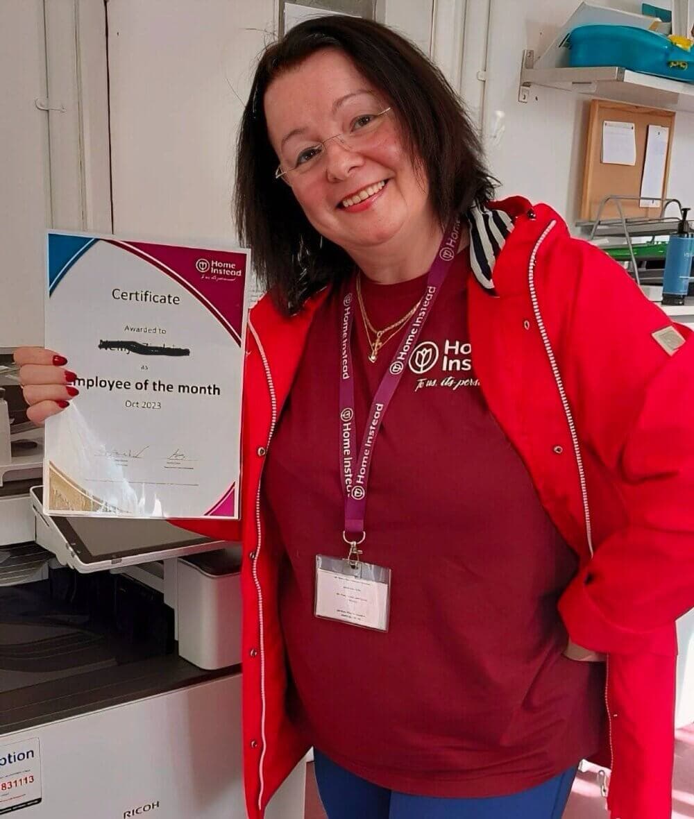 A woman in a red jacket smiles while holding an "Employee of the Month" certificate in an office. - Home Instead