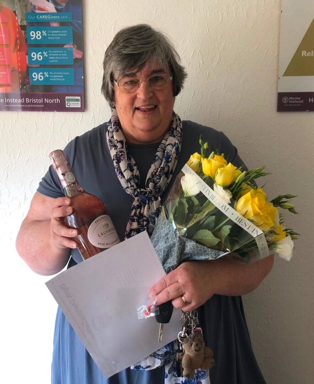 A woman smiles while holding a bouquet of yellow flowers, a bottle of rosé wine, and a set of keys. - Home Instead