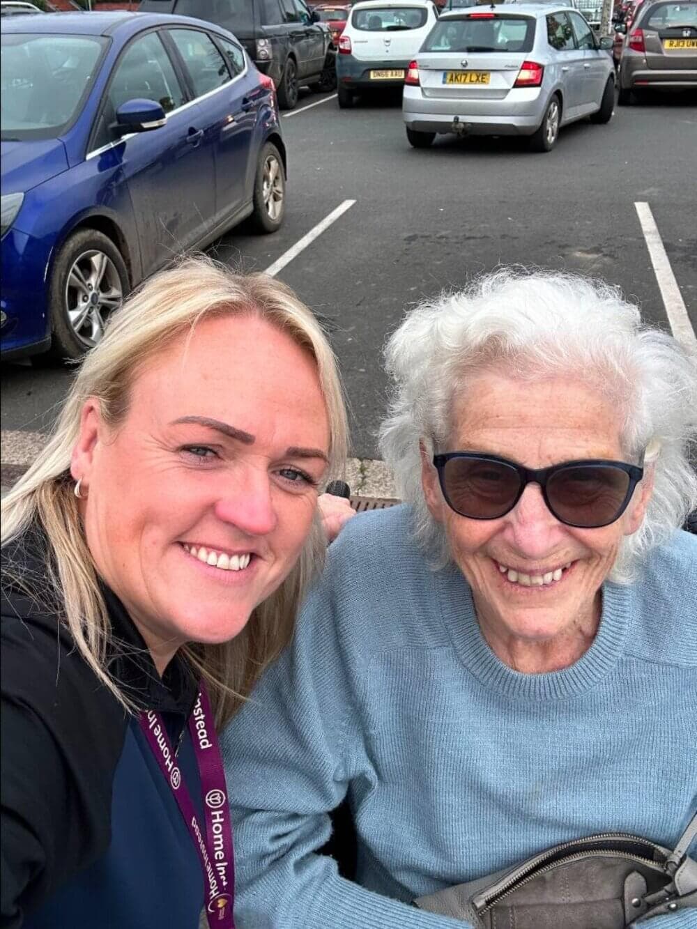 A smiling woman and elderly woman in sunglasses take a selfie in a parking lot with cars in the background. - Home Instead