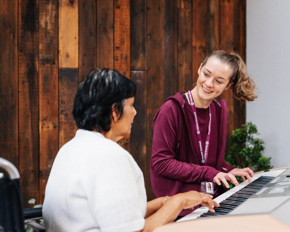 A woman teaches another woman to play the keyboard, both smiling in a warmly lit room with wooden walls. - Home Instead