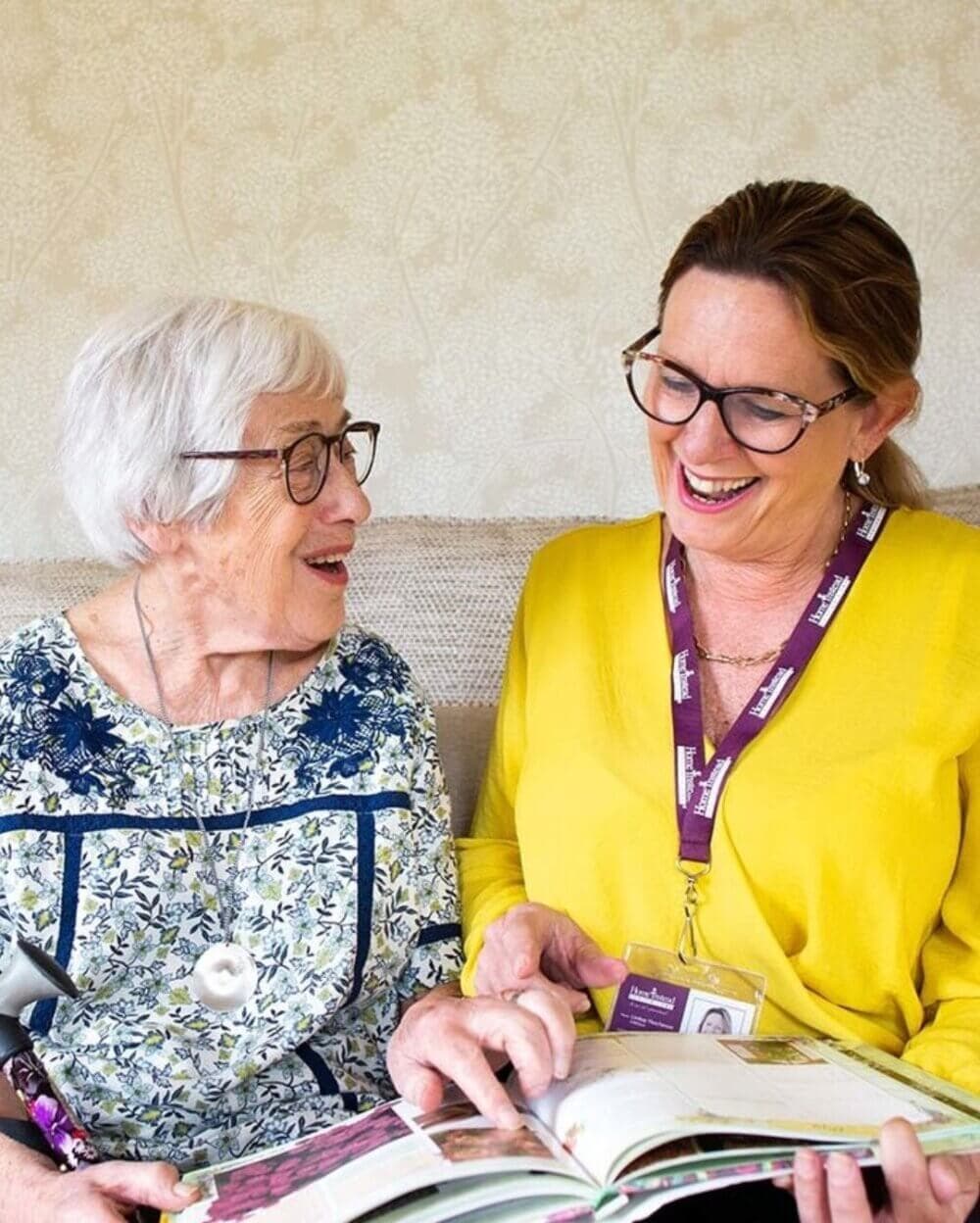 An elderly woman and a caregiver share a smile while looking at a book together. - Home Instead