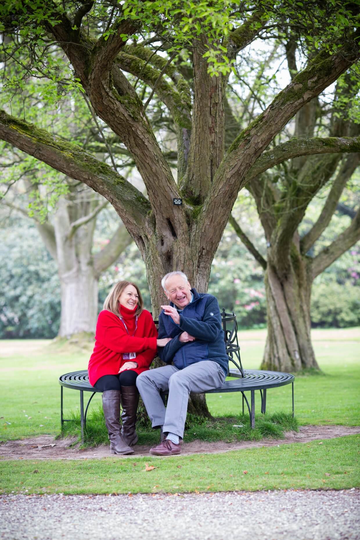 An elderly couple sitting on a circular bench around a tree, laughing and enjoying each other's company in a park. - Home Instead