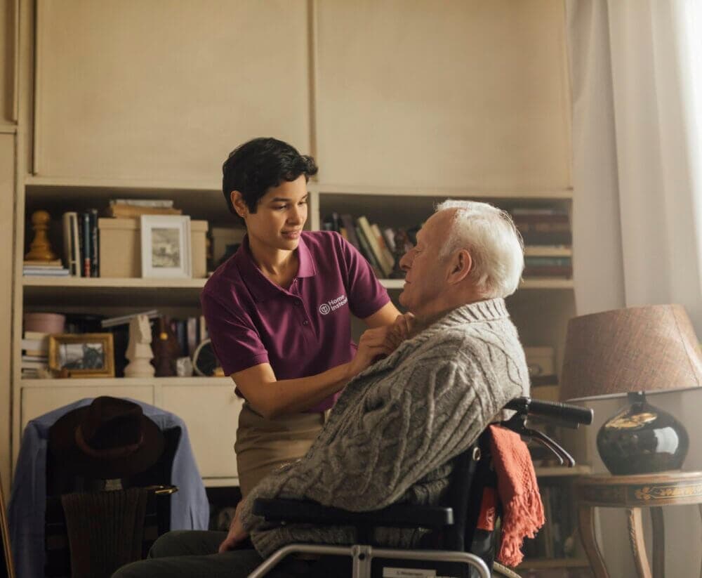 A caregiver in a purple shirt helps an elderly man in a wheelchair adjust his sweater in a cozy, book-filled room. - Home Instead