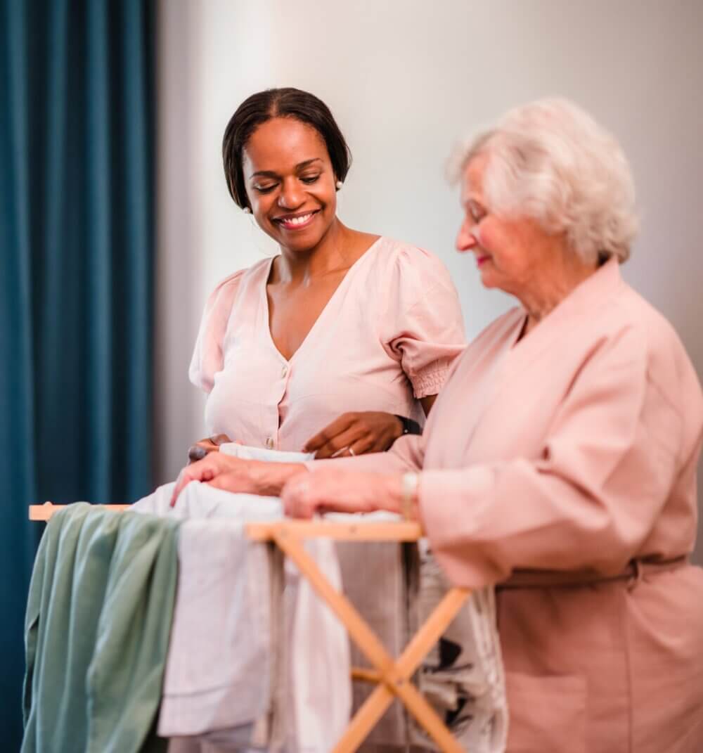 Two women, one younger, one older, folding clothes together while smiling and conversing in a warmly lit room. - Home Instead