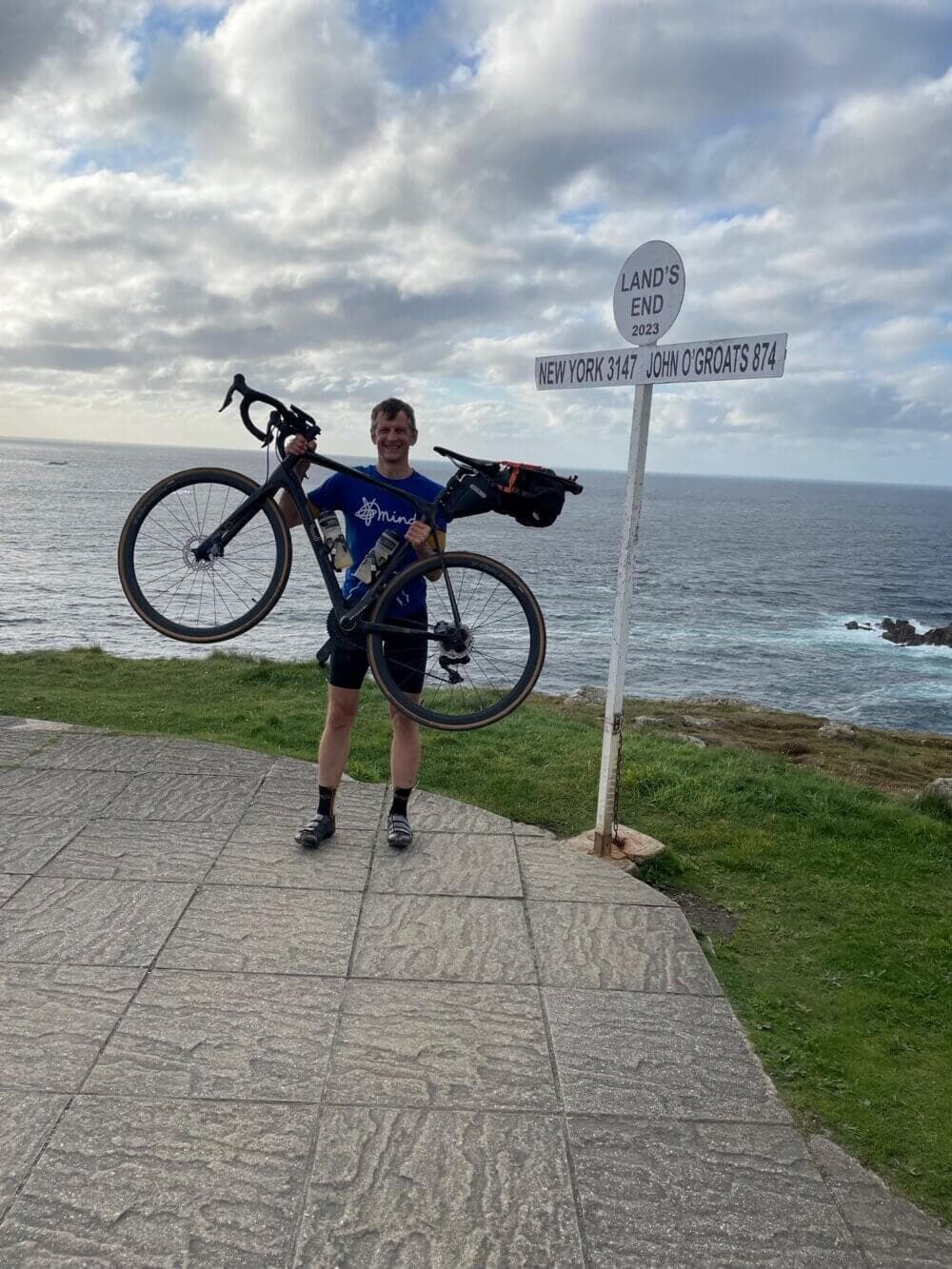 Person holding a bicycle overhead at Land's End with sign showing distances to New York and John O'Groats, near the sea. - Home Instead