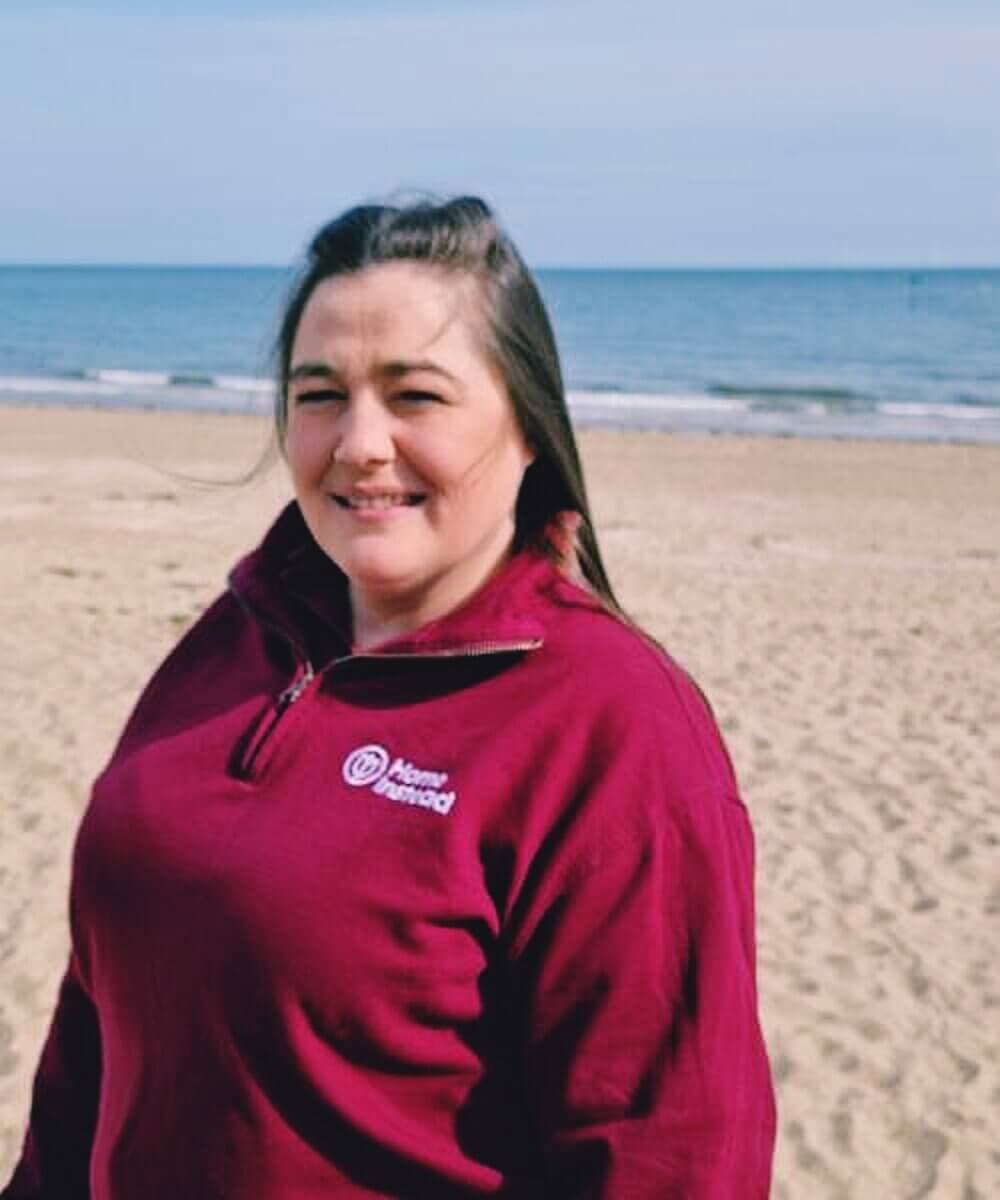 Person in a red sweater standing on a beach with the ocean in the background on a clear day. - Home Instead