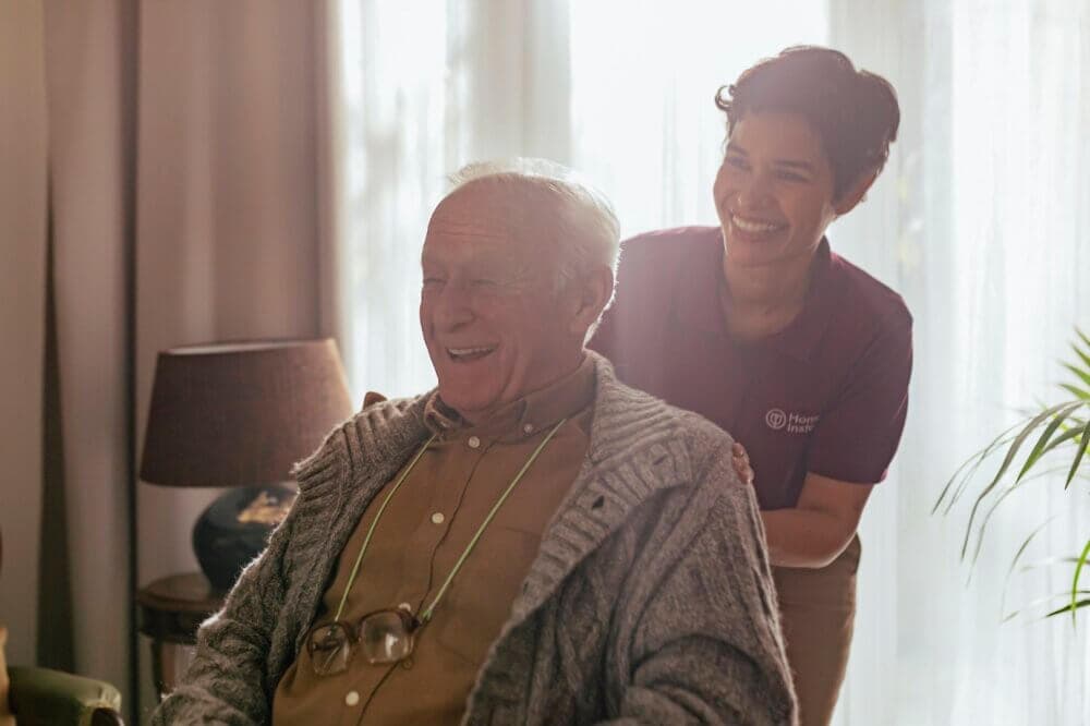 An elderly man smiles while seated, with a caregiver standing behind him, both sharing a light moment in a sunlit room. - Home Instead
