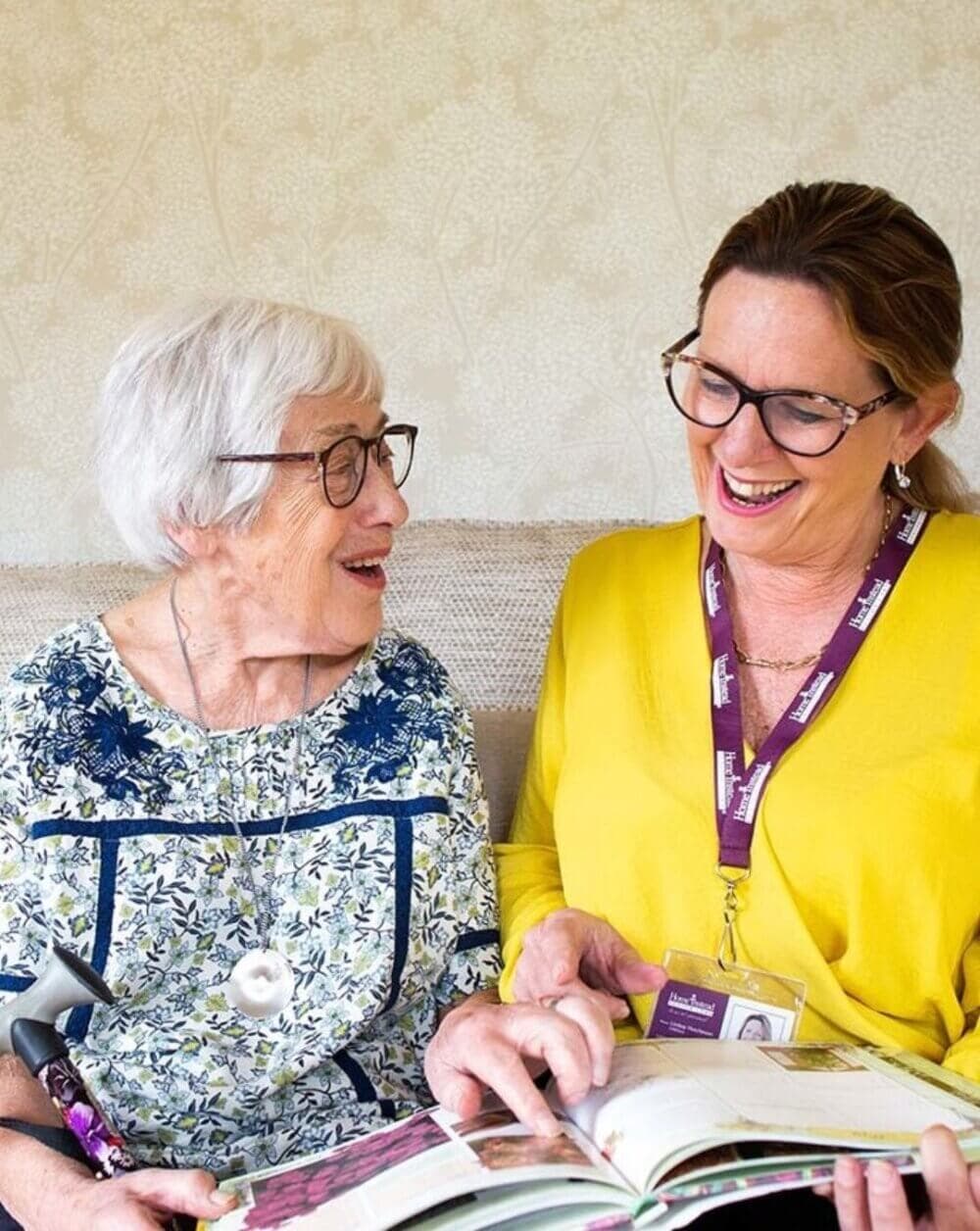 An elderly woman and a caregiver share a laugh while looking at a book together on a couch. - Home Instead