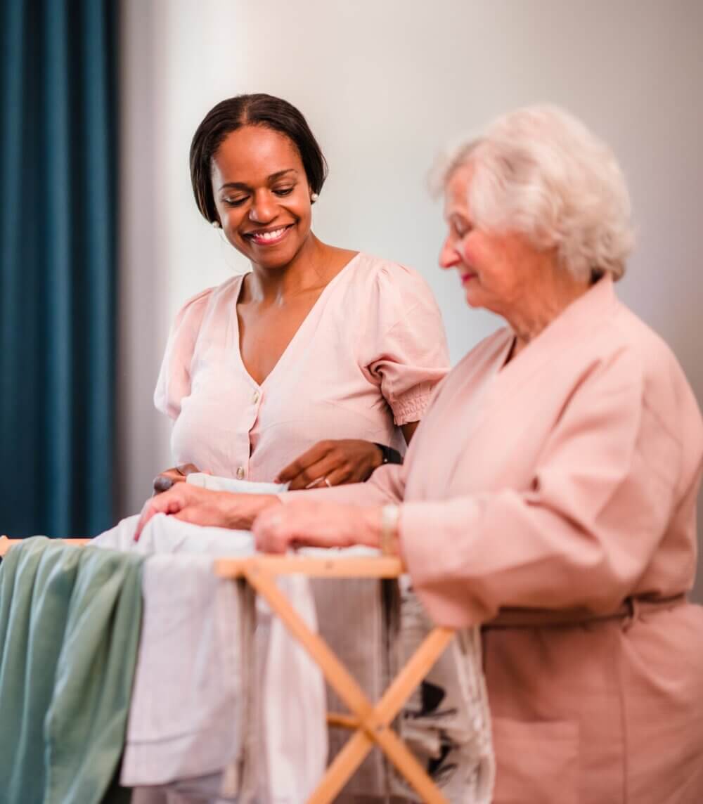 Two women smiling and folding laundry together, one younger with short hair and the other older with gray hair. - Home Instead