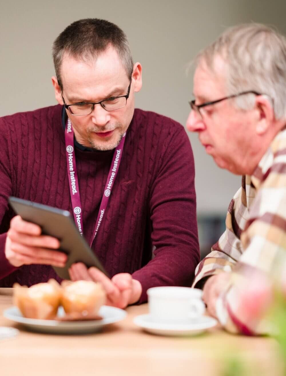 Two men focus on a tablet over coffee and muffins; one seems to be explaining something on the device to the other. - Home Instead