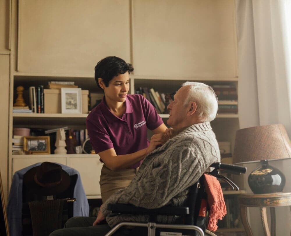 Caregiver in a purple shirt gently assists an elderly man in a wheelchair in a cozy, book-filled room. - Home Instead
