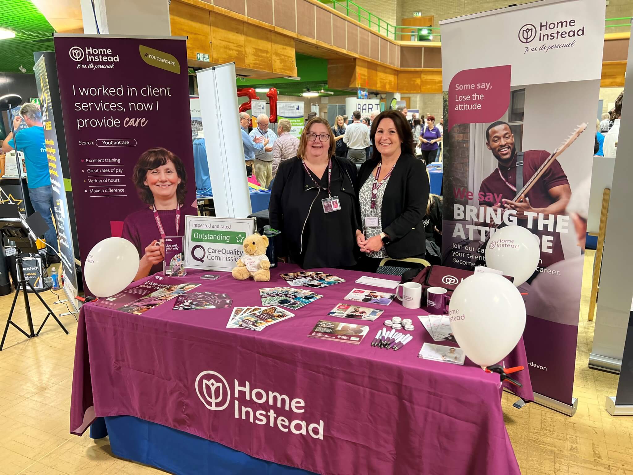 Three women at a Home Instead booth with promotional materials and balloons at a community event. - Home Instead