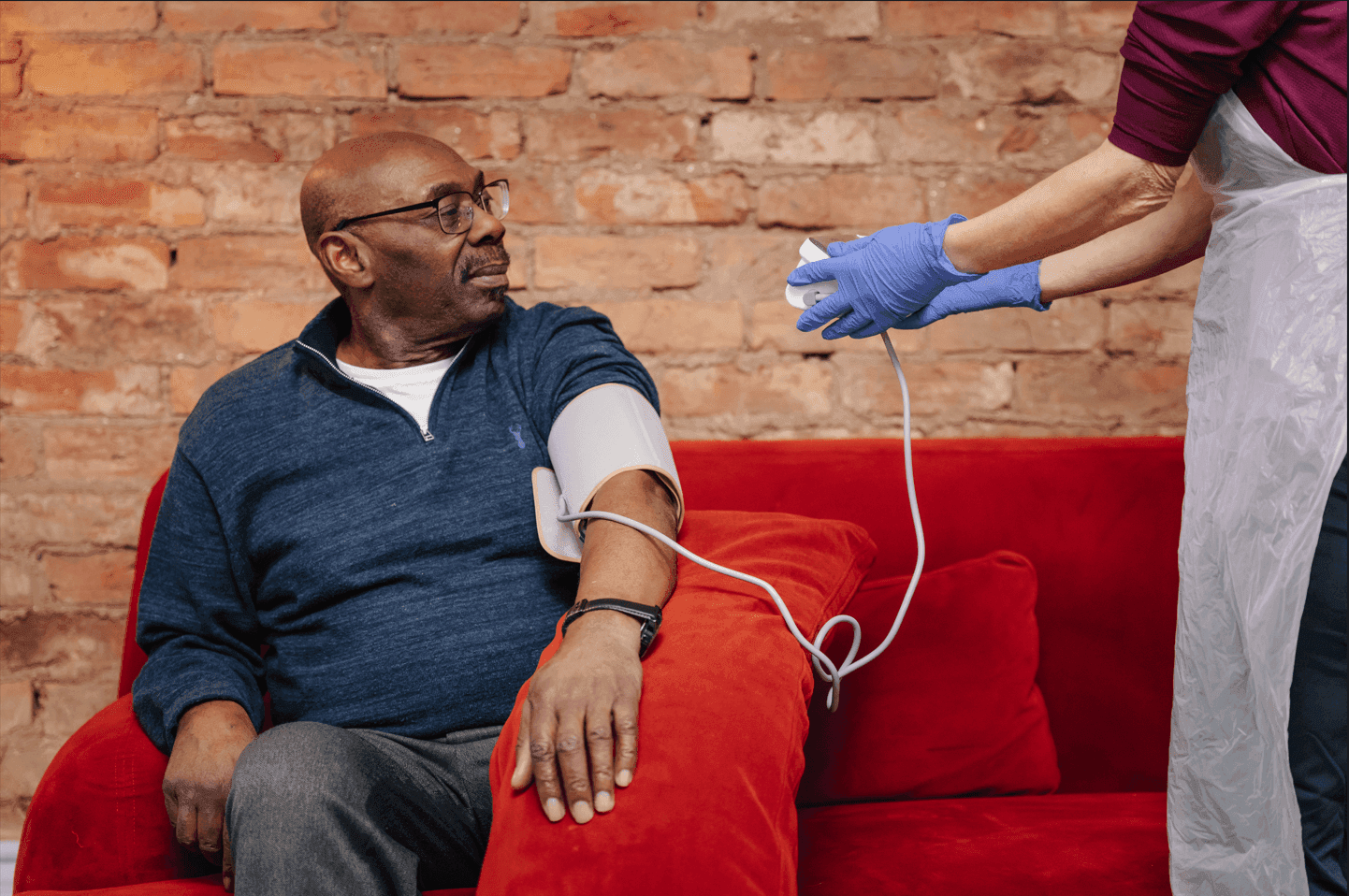 A man sits on a red sofa getting his blood pressure checked by a healthcare worker in a blue glove. - Home Instead