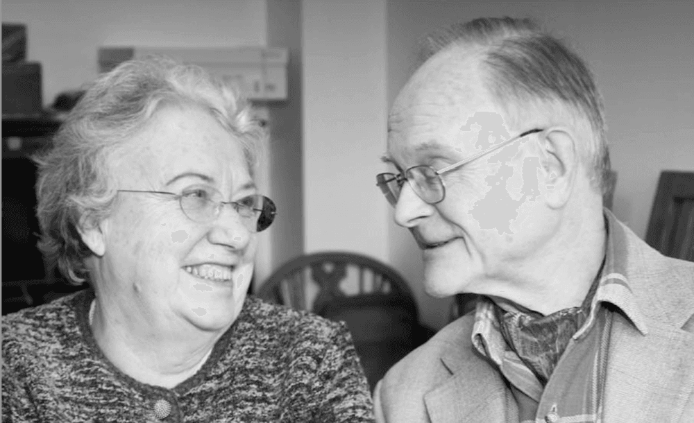 An elderly couple with glasses smile warmly at each other in a black and white photo. - Home Instead