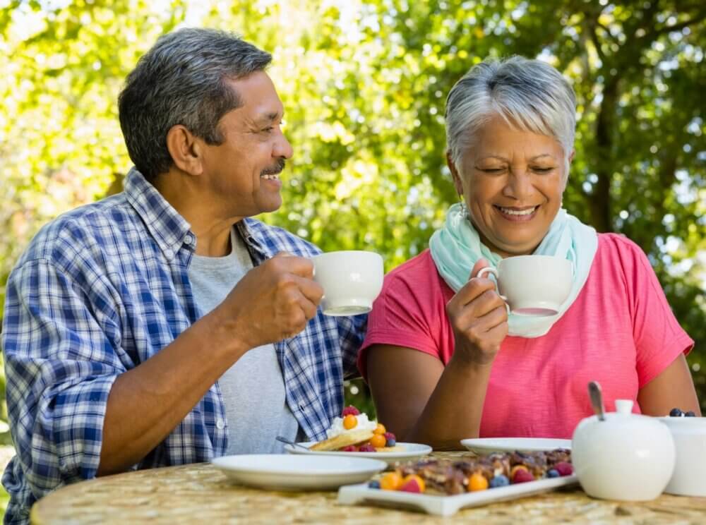 An elderly couple sits outdoors, smiling and drinking tea or coffee together at a table. - Home Instead