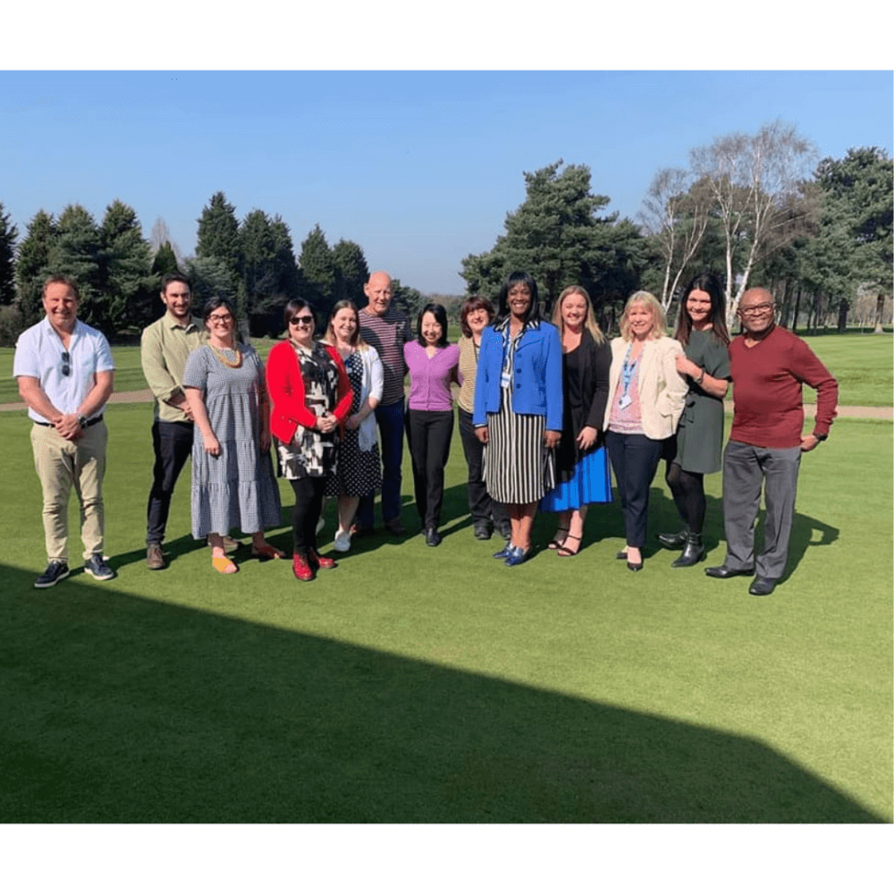 A diverse group of 13 adults stands on a grassy field with trees in the background, smiling at the camera. - Home Instead