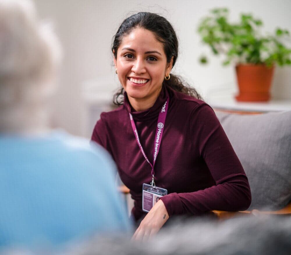 A woman wearing a name badge smiles while talking to another person in a cozy room with a potted plant in the background. - Home Instead