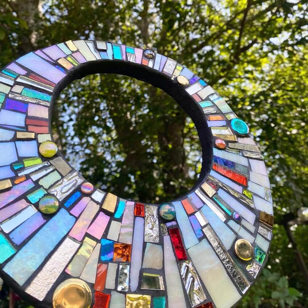 A colorful circular mosaic art piece set against a backdrop of leafy green trees. - Home Instead