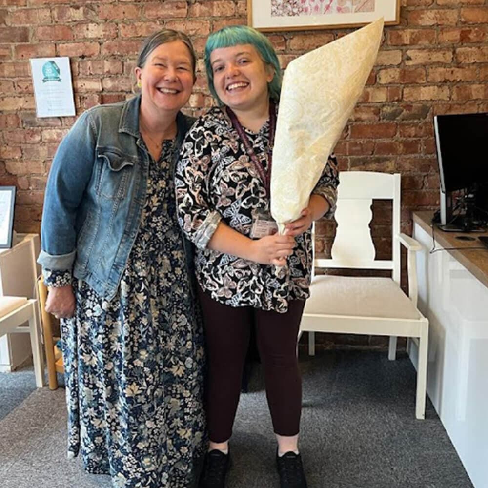 Two smiling people pose together; one holds a bouquet wrapped in paper. Brick wall and office furniture in the background. - Home Instead
